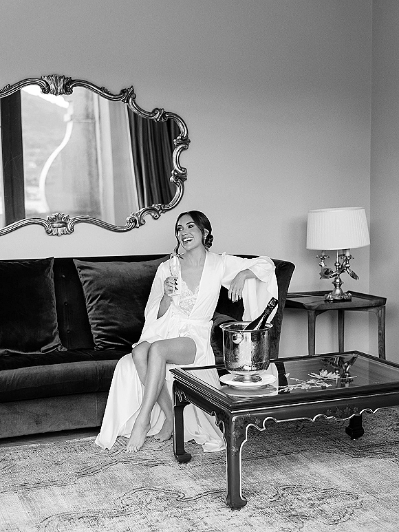 Bridal portrait of a bride getting ready in a silk robe and lingerie, holding champagne beside an ornate mirror in a hotel suite