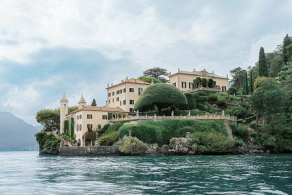 Wedding venue exterior with a historic villa on a stone terrace, framed by topiary gardens, overlooking a lake and mountains under clouds