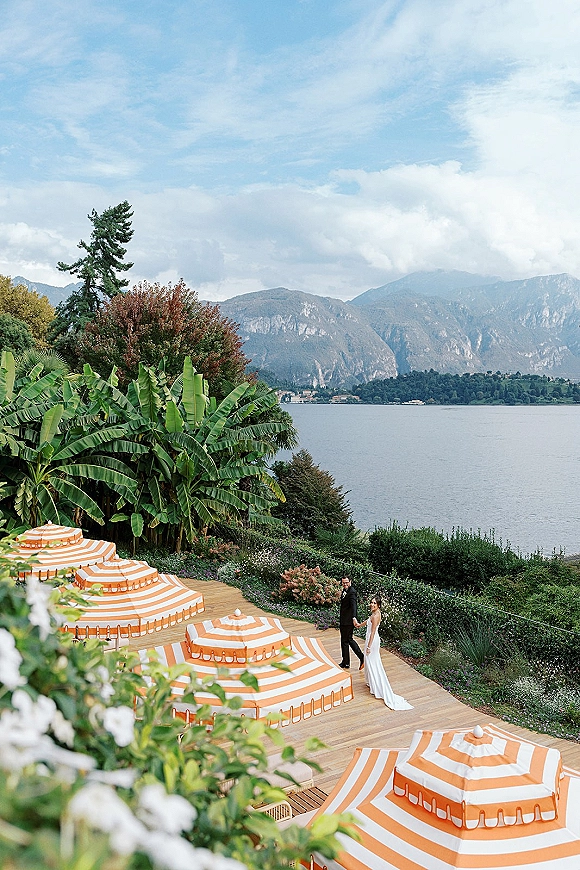 Couple portrait of bride and groom walking hand in hand on a wooden deck with striped umbrellas, with a mountain lake backdrop