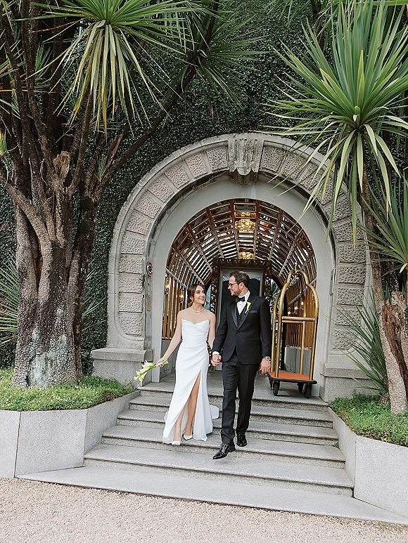 Couple portrait of newlyweds walking together, holding hands on steps under a stone archway, bride with calla lily bouquet and strapless gown