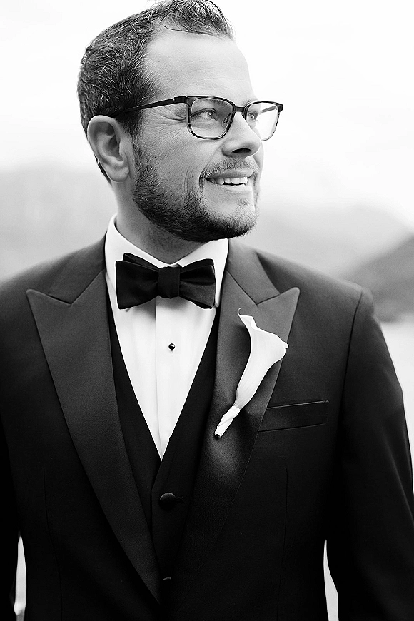 Groom portrait in a black tuxedo with bow tie and eyeglasses, boutonniere on lapel, standing outdoors against an open sky backdrop