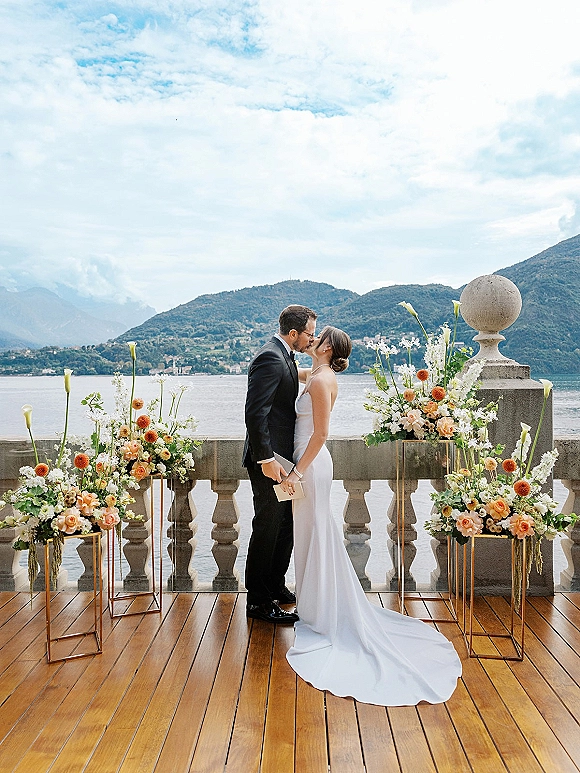 Wedding kiss portrait of bride and groom kissing on a wooden deck by a lakeside with mountains, framed by peach roses on gold stands