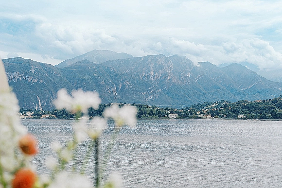 Wedding landscape photo with out-of-focus flowers in the foreground, overlooking a mountain lake, shoreline village, hills, and cloudy sky