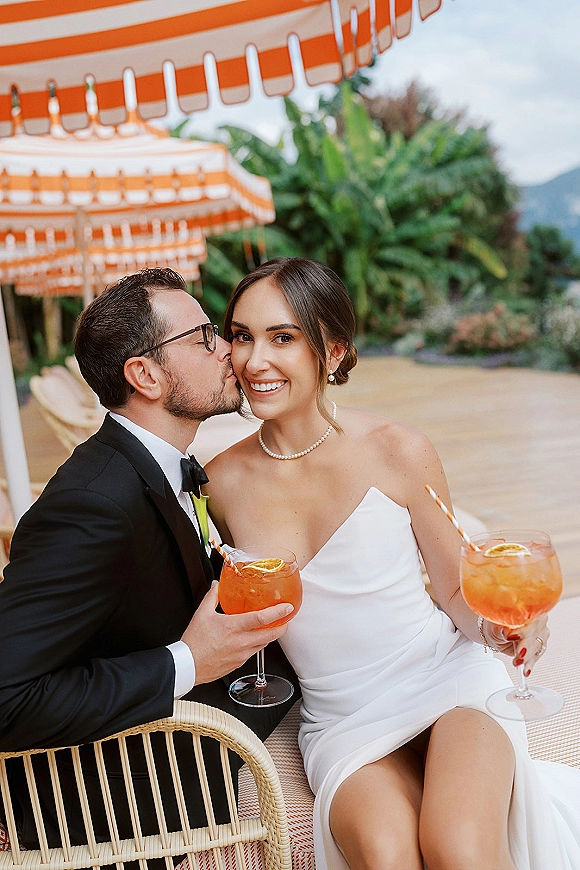 Couple portrait of bride and groom drinks as he kisses her cheek, toasting with citrus cocktails under striped umbrellas on a patio.