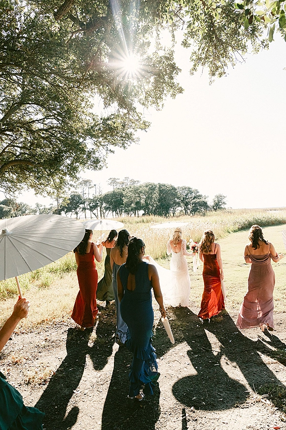 Wedding party walking as the bride walks with bridesmaids behind her, bouquet in hand, parasol umbrellas under a sunlit tree canopy