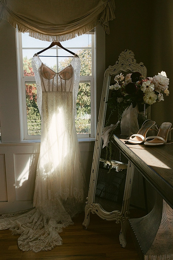 Wedding dress with lace wedding dress details hanging by a sunlit window, long train draped near bouquet, white heels, and ornate mirror