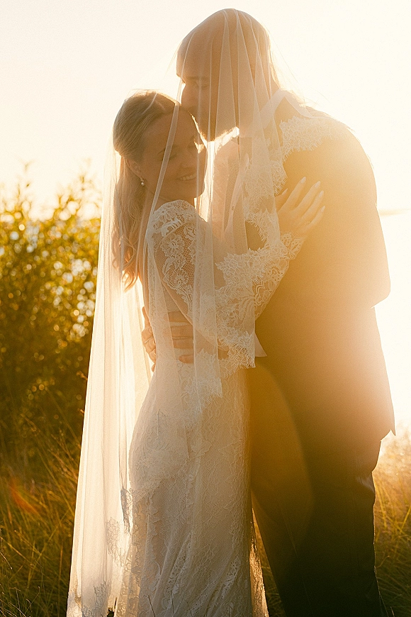 Wedding couple portrait of bride and groom embrace as he kisses her forehead, long veil over them in golden sunset light among tall grass