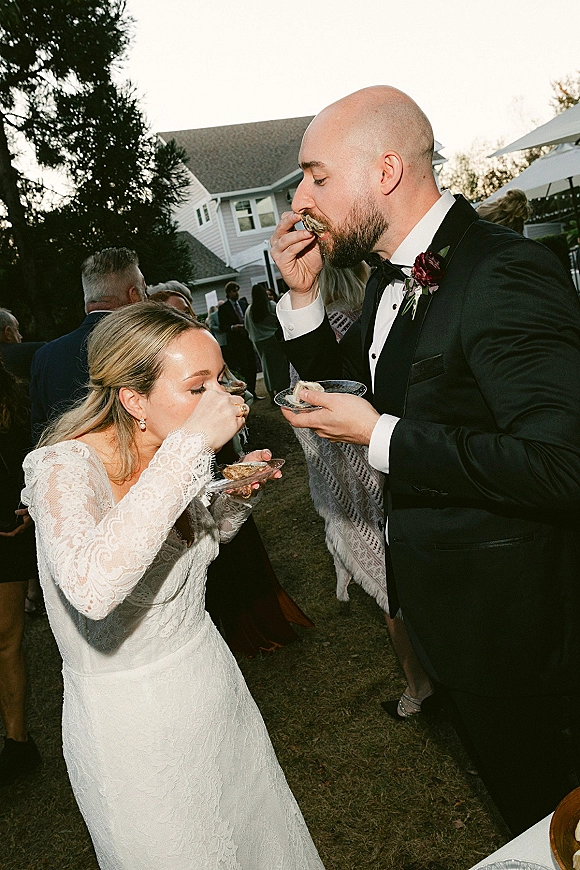 Wedding reception moment of bride and groom eating oysters at a cocktail table, her lace long sleeves and his tuxedo with burgundy boutonniere on a lawn under a tent