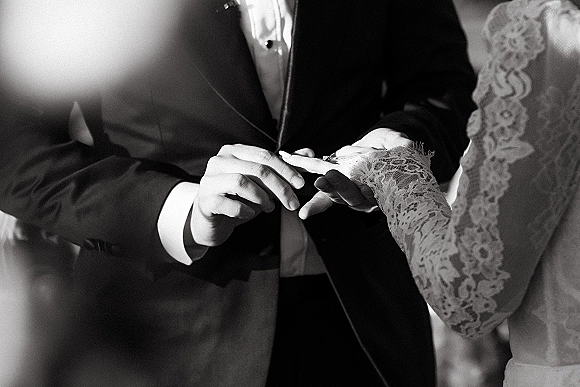 Wedding ring exchange as groom places a band on bride’s hand, lace sleeves and tuxedo cufflinks in soft-lit indoor ceremony