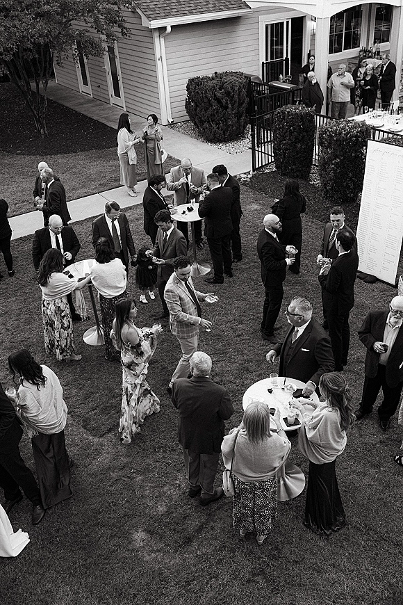 Cocktail hour with wedding guests mingling around high top tables, holding drinks and chatting in suits and dresses on a lawn patio