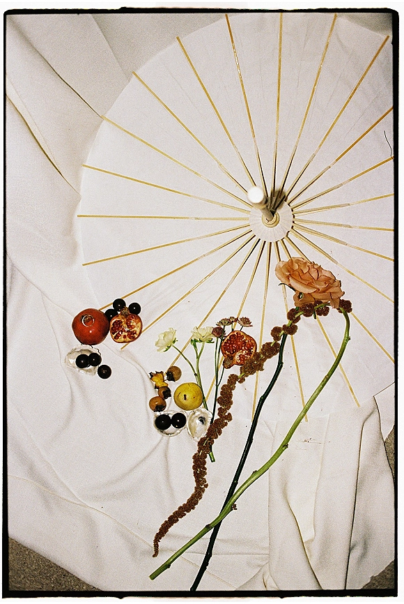 Wedding flat lay bridal detail flat lay with white paper parasol, rose, greenery, pomegranate, grapes, citrus, and oysters on white fabric backdrop