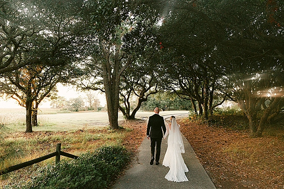 Couple walking away, bride and groom walking hand in hand down a tree-lined oak path with sunlight rays and her veil trailing behind