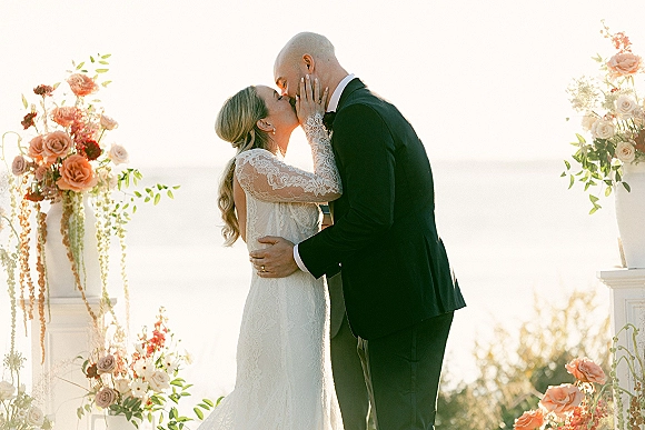 Wedding kiss portrait of bride and groom kissing, bride in lace long sleeves holding his face by floral pillars with ocean horizon behind