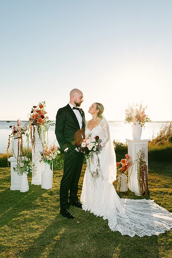 Couple portrait of bride and groom portrait gazing at each other, bride holding bouquet with long veil by a waterfront at sunset.