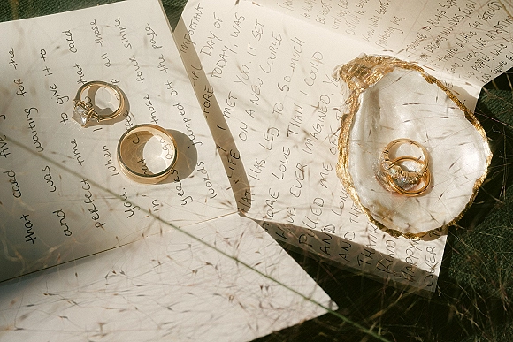 Wedding rings and diamond engagement ring in an oyster shell dish over handwritten letters on sunlit grass with soft shadows