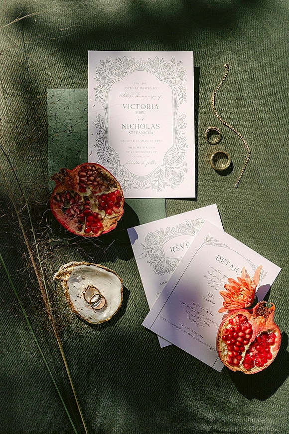 Invitation suite wedding invitation flat lay on green linen with RSVP and details cards, rings in an oyster shell, pomegranate and dried grass