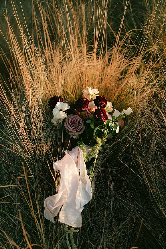 Bridal bouquet with burgundy and white bouquet roses and greenery tied with a white ribbon, resting in tall grass in a field