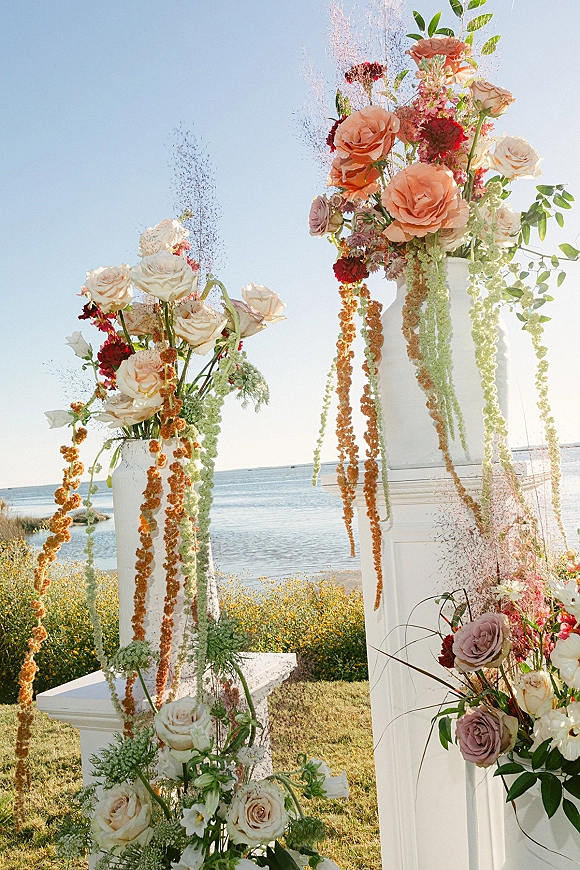 Ceremony aisle decor with wedding aisle flowers on white pedestals, rose and greenery pillars, hanging strands, set by the ocean shoreline