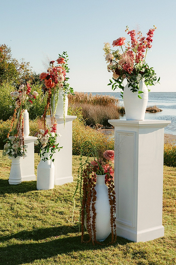 Ceremony altar decor with wedding altar flowers in white vases and pedestals, roses and trailing greenery, set by a waterfront lawn under blue sky