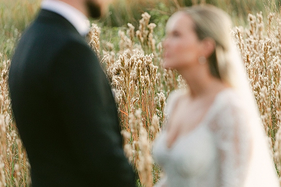 Wedding ceremony moment as bride in a long veil and sheer-sleeve gown faces groom in a black tux in a tall grass field, golden hour