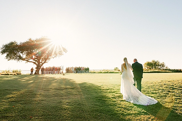 Wedding processional with bride walking down aisle in lace dress and veil, bouquet in hand, on grassy lawn by tree and water horizon at sunset