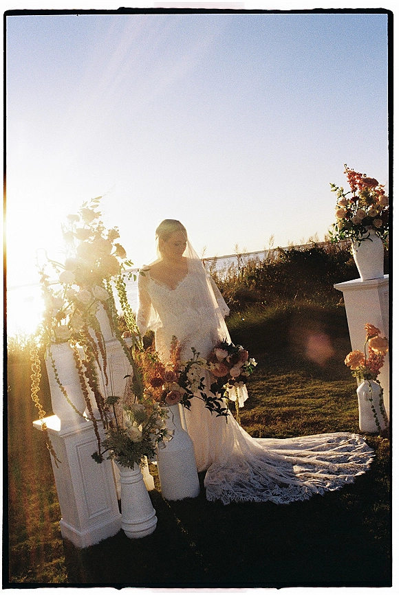 Bridal portrait of a bride with veil in a lace wedding dress train, standing by floral columns on a lawn near the shoreline at sunset