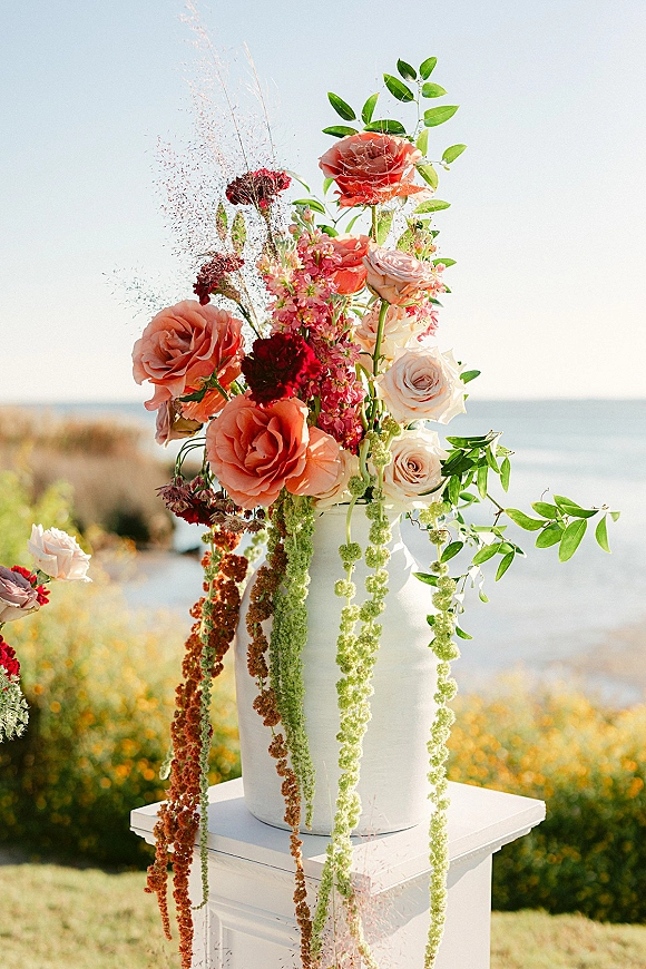 Wedding floral arrangement of roses and greenery with hanging amaranthus in a ceramic vase on a pedestal, set against an ocean coastline backdrop