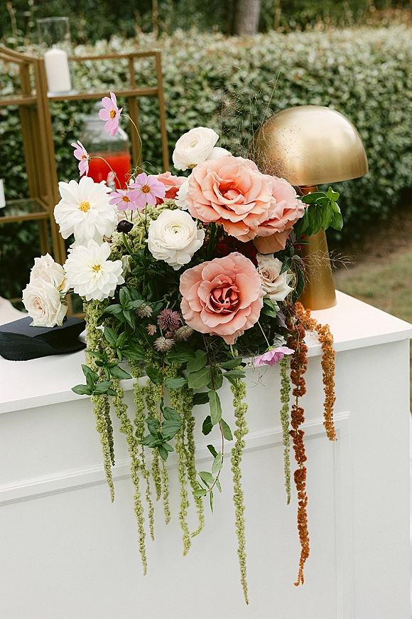Wedding floral arrangement with blush roses, dahlias, and greenery cascading from a white pedestal table in a garden hedge setting