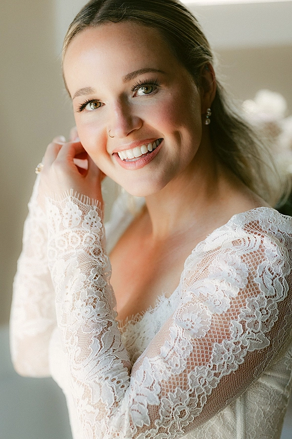 Bridal portrait of a smiling bride in a lace wedding dress with long sleeves and drop earrings, softly lit by window light indoors