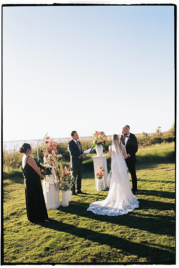 Wedding vows as bride and groom stand at an outdoor wedding ceremony under a floral arch on a coastal lawn with ocean horizon backdrop