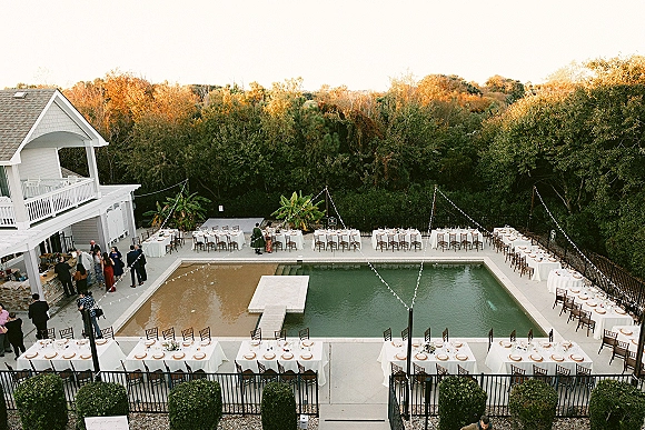 Poolside reception setup with long white-linen banquet tables, wooden chairs, greenery centerpieces, and string lights around a sunset pool deck