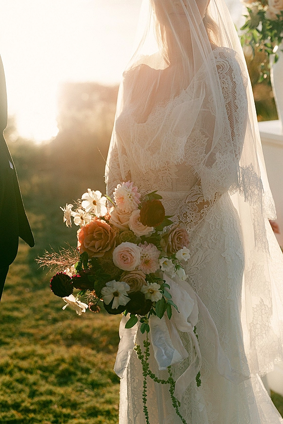 Bride portrait backlit bridal portrait with lace dress and cathedral veil, holding rose and dahlia bouquet with ribbon in a sunset field