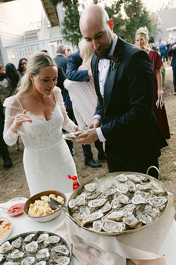 Wedding cocktail hour oyster bar wedding scene with bride in lace dress and groom in tuxedo beside oysters on ice at lawn reception