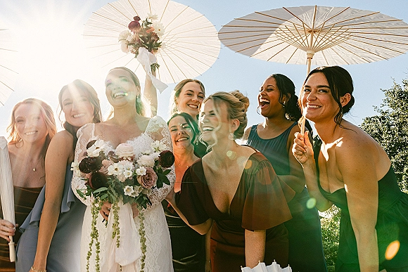 Bridesmaid group photo with bride with bridesmaids holding paper parasols, bride in lace dress with bouquet under blue sky and trees