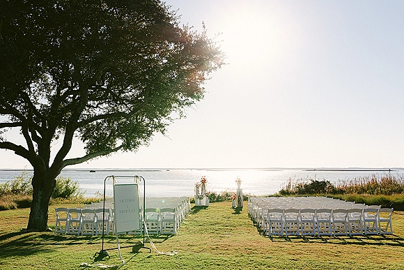 Ceremony setup with white folding chairs and aisle florals on a grassy waterfront lawn, welcome sign and ribbon streamers in sun flare