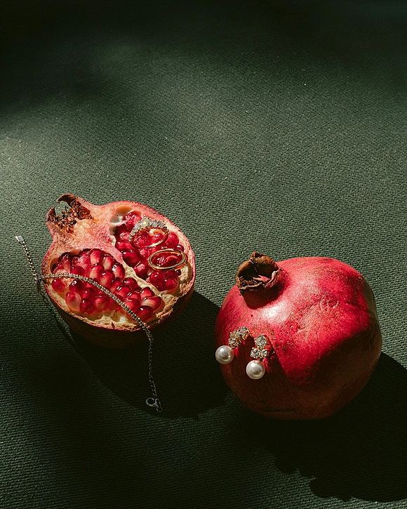 Wedding rings ring flat lay with gold bands and a diamond ring alongside pearl drop earrings, tennis bracelet, and pomegranate on green fabric