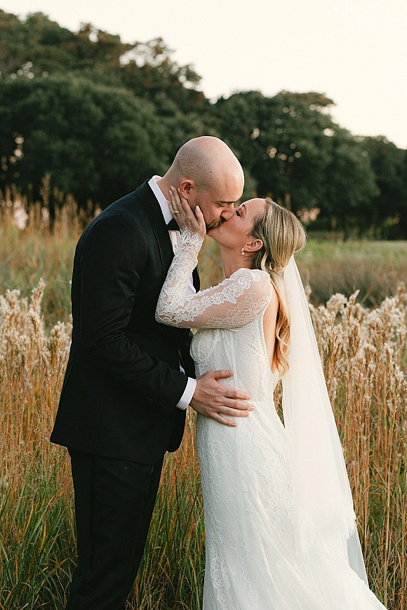Wedding kiss portrait of bride and groom kissing in a tall grass field at golden hour, bride in lace long sleeves with veil and pearls