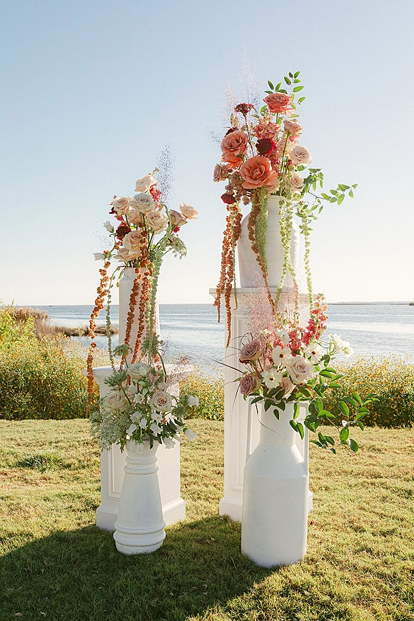 Ceremony floral pillars on white pedestals with roses and hanging amaranthus, set on a lawn with ocean horizon backdrop