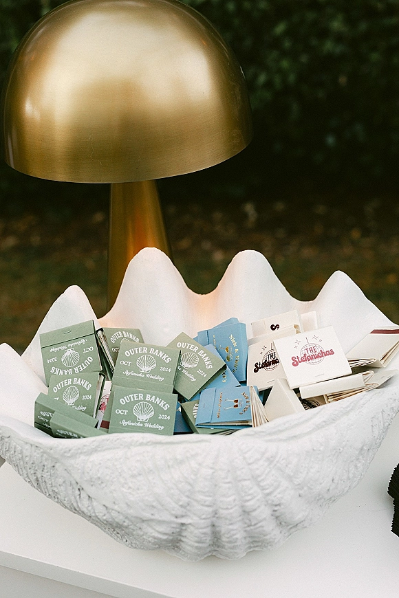 Wedding matchbooks displayed in a scallop shell bowl on a tabletop with green foliage and a brass lamp, custom matchbooks wedding favor detail