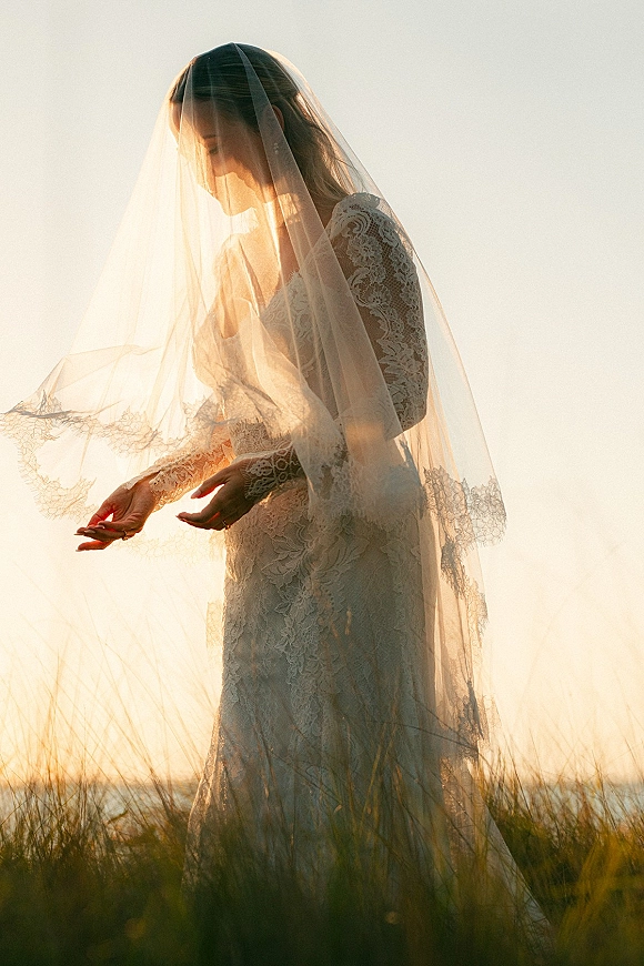 Bridal portrait of a bride silhouetted at sunset, cathedral veil over her face, lace long sleeves, standing in tall grass by water horizon