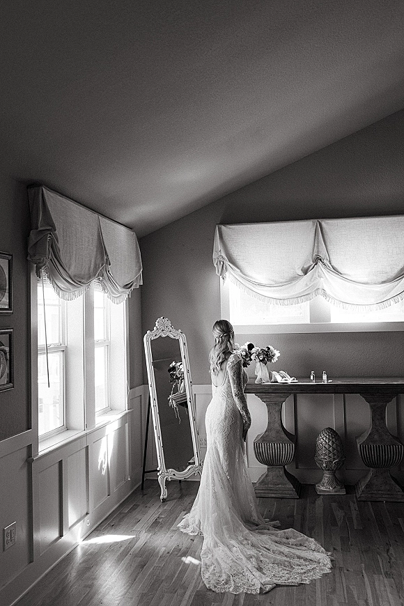 Bridal portrait of a bride in a lace wedding dress with a long train holding a bouquet by large windows, beside a floor mirror in soft light