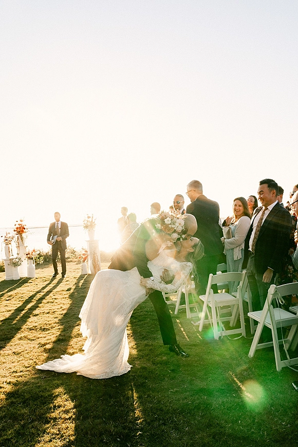 Wedding kiss portrait at sunset as groom dips bride in lace-sleeve gown holding bouquet, with waterfront lawn ceremony chairs behind