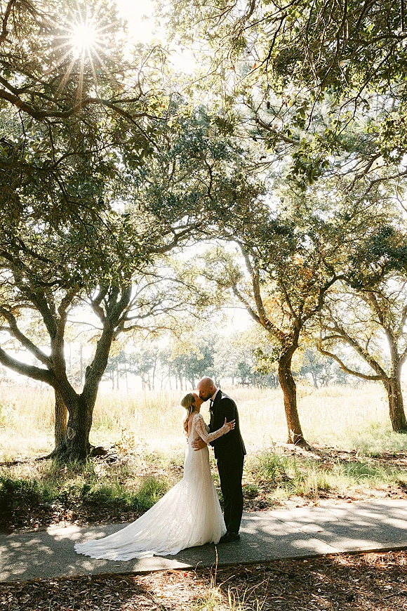 Wedding kiss portrait of bride and groom kissing at an outdoor wedding kiss, bride in lace long sleeves with sun flare under oak trees