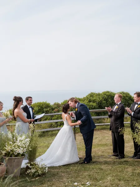 Ceremony kiss as bride and groom hold hands by oceanfront wedding ceremony florals, with wedding party on a lawn beside a wooden fence