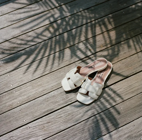 Bridal shoes, white wedding sandals with square toes on a sunlit wood deck floor, casting crisp shadow patterns