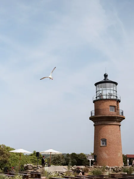 Outdoor wedding ceremony with rows of chairs and white umbrellas set beside a brick lighthouse under blue sky, with a seagull overhead