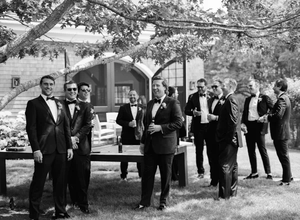 Groomsmen candid in tuxedos and bow ties lounging in outdoor chairs with beer bottles, laughing on a lawn by a house porch