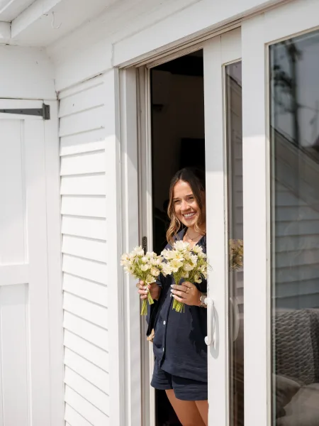 Bridesmaid portrait of a woman holding white flower bouquets in matching pajama set by a sliding glass door with window reflections