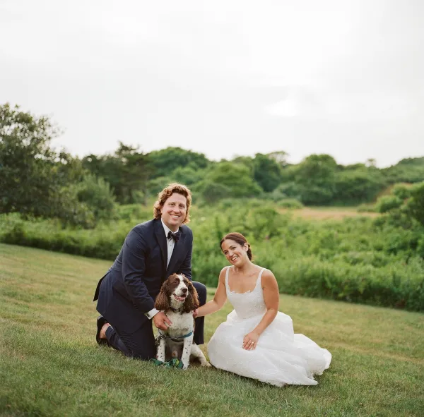 Couple portrait of bride and groom with dog, kneeling on grass with leash, her wedding dress and his tuxedo in green hills