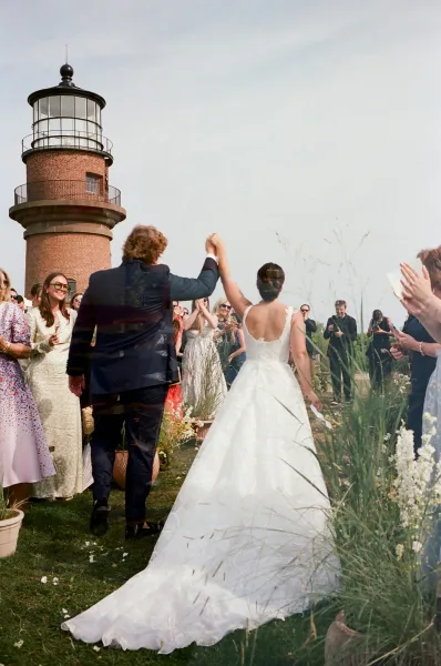 Wedding recessional as newlyweds walk back up aisle with raised hands, guests cheering on a lawn with wildflowers near a lighthouse
